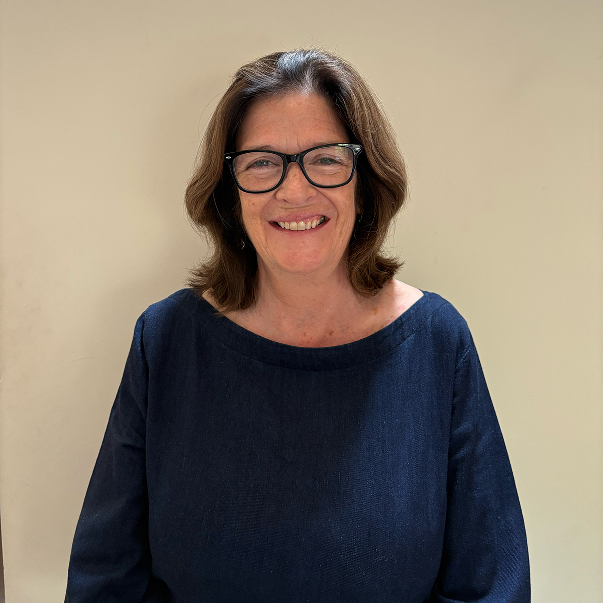 Head-and-shoulders portrait of an adult wearing glasses and a dark top, standing against a plain light-coloured background.