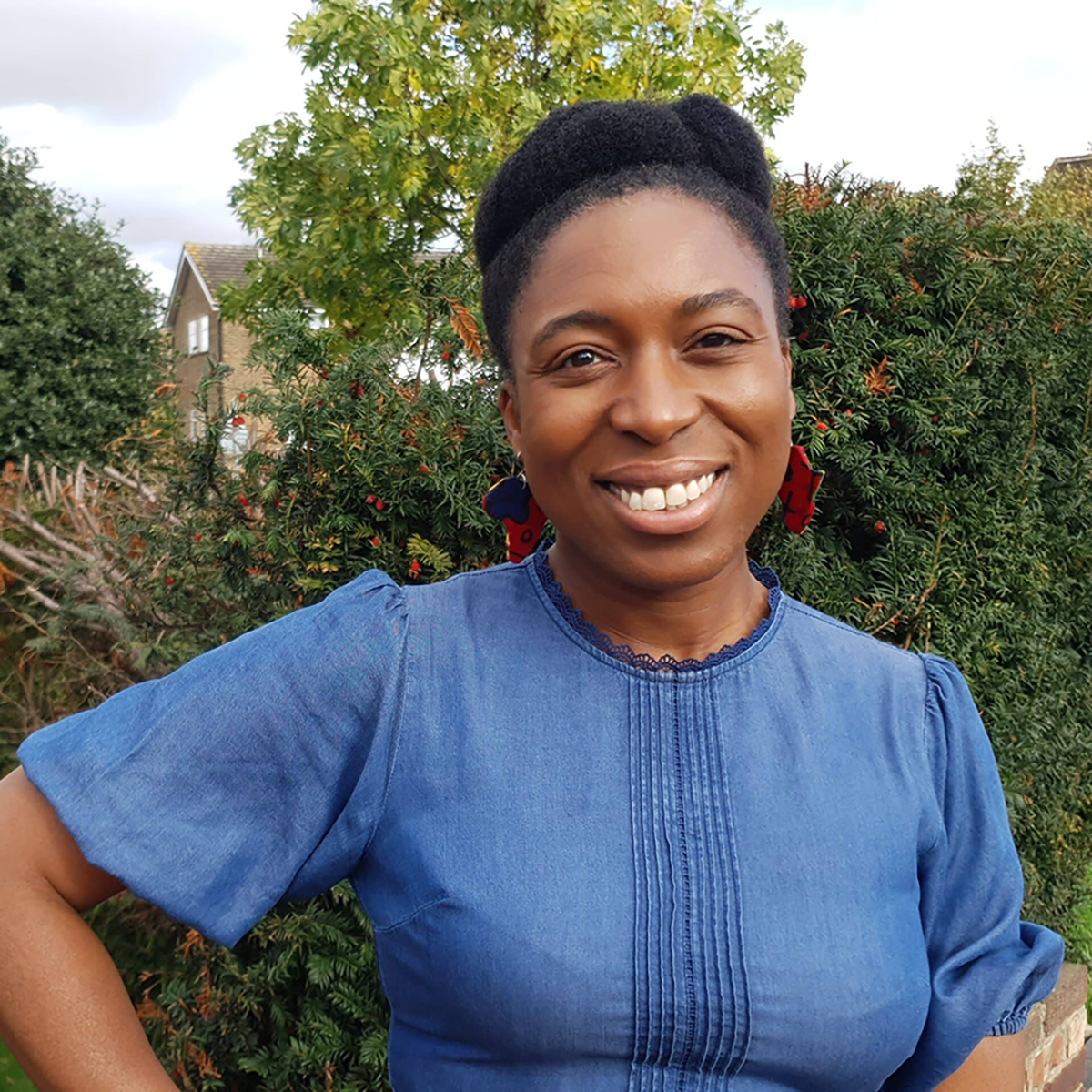 Portrait of an adult wearing a blue top and statement earrings, standing outdoors in front of green foliage and residential houses.