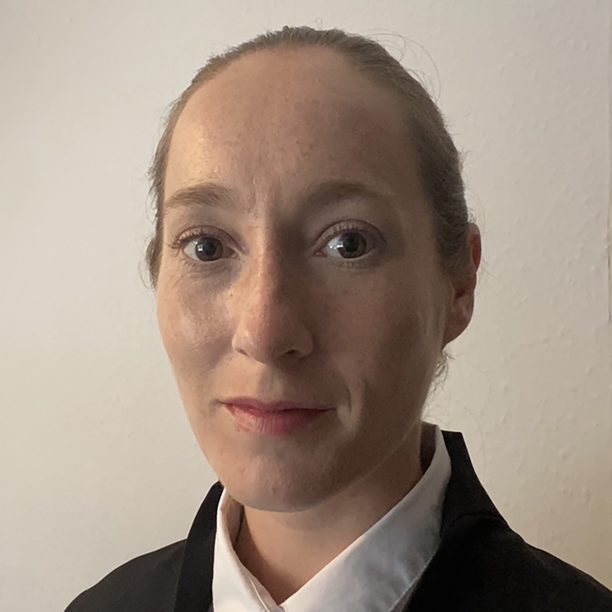 Close-up portrait of an adult with hair pulled back, wearing a dark jacket and light shirt, against a plain light background.