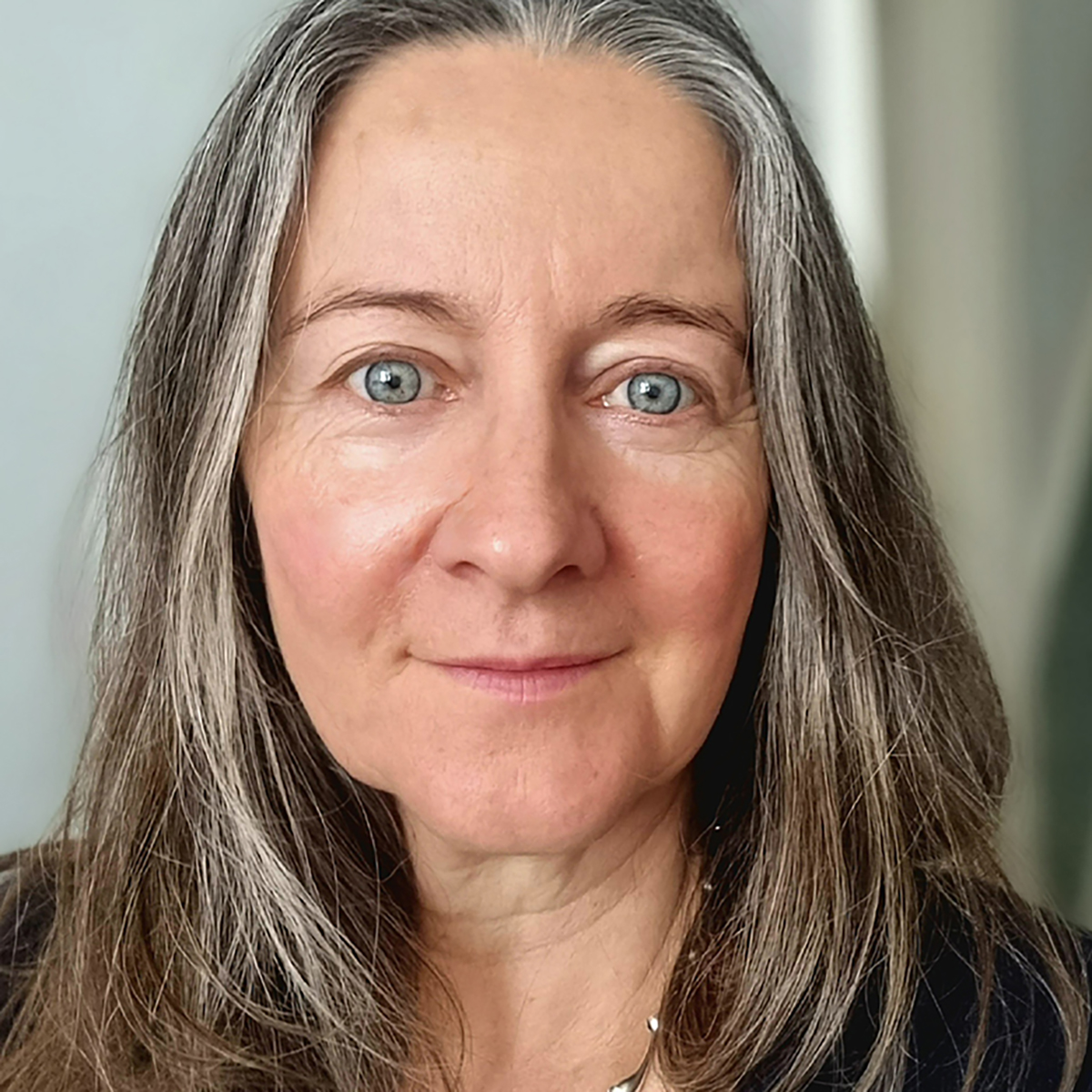 Head-and-shoulders portrait of an adult with long grey hair, wearing a dark top, facing the camera against a softly blurred indoor background.