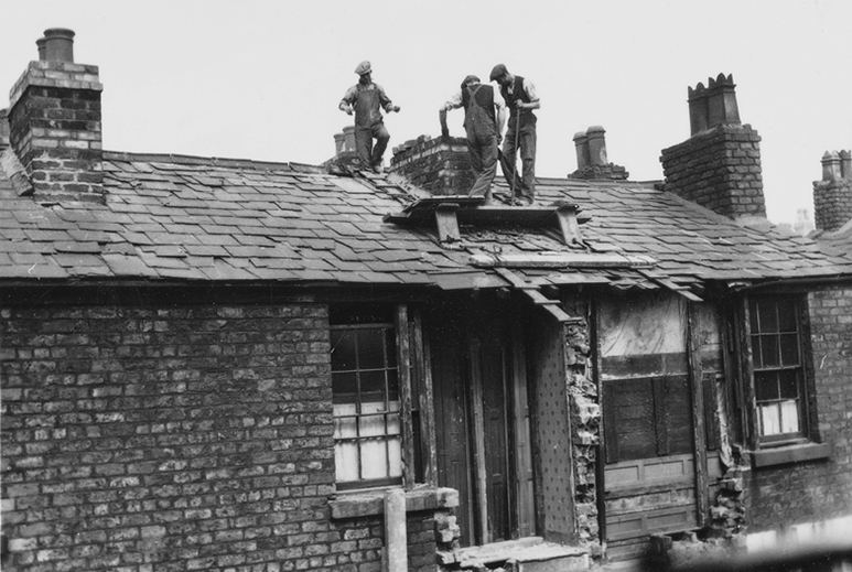 Black-and-white photo of several workers standing on the roof of a brick house, carrying out repairs around a damaged section with exposed structure, chimneys visible along the roofline.