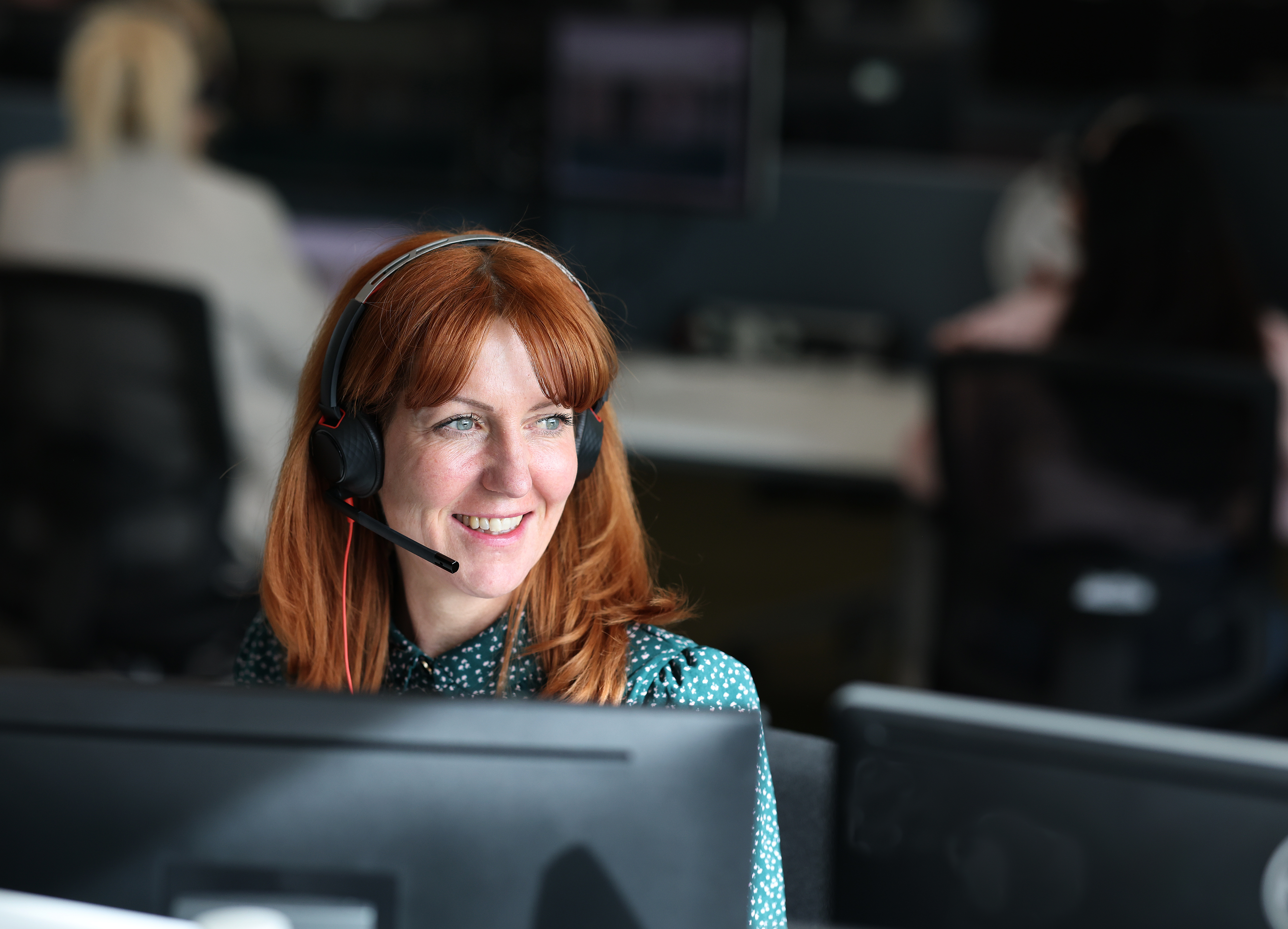 Head-and-shoulders view of an adult wearing a headset and microphone, seated at a computer in an office, with blurred colleagues and desks in the background.