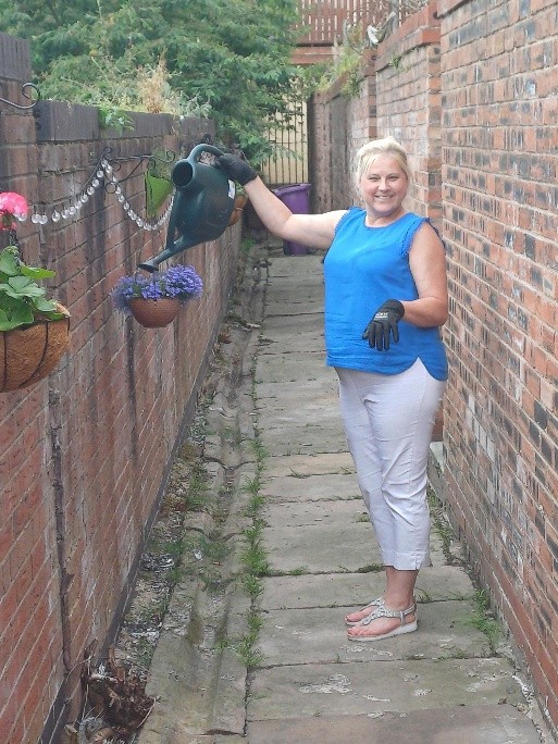 Person wearing a blue sleeveless top, light trousers and gardening gloves, watering hanging plants along a narrow paved alley bordered by red-brick walls.