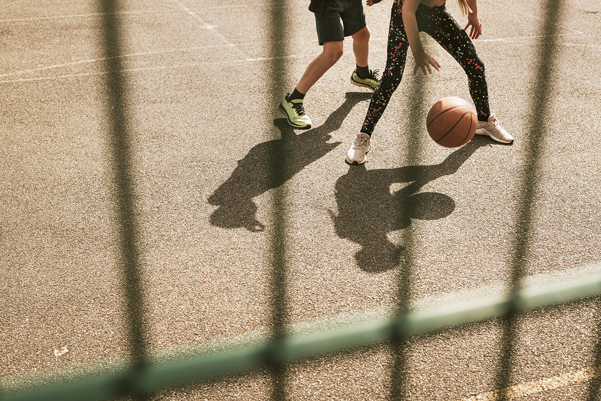 Two people playing basketball on an outdoor court, with a ball mid-bounce and shadows visible through a metal fence in the foreground.